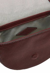 Sac en cuir bordeaux avec surface texturée, surpiqûres rouges et intérieur lisse. À l'intérieur, on trouve une étiquette embossée avec "Fossil."