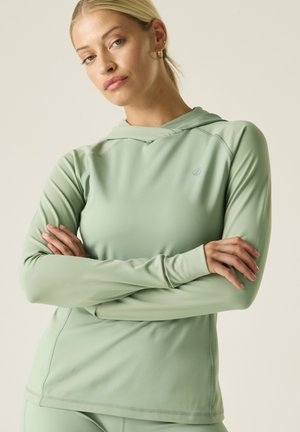 Blonde Frau mit verschränkten Armen, die ein hellgrünes langärmeliges Hoodie-Shirt mit kleinem Logo auf der Brust vor einfarbigem Hintergrund trägt.