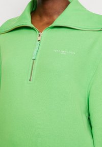 Zelená mikina se zipem vyrobená z pleteného materiálu s vysokým límcem, s kovovým zipem a malým bílým logem na hrudi.