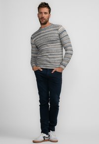 Pull rayé gris clair avec des accents bleus et bruns, assorti à un pantalon bleu foncé et des baskets blanches avec des semelles brunes.