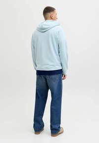 Homme aux cheveux courts portant un sweat à capuche bleu clair, une chemise bleu foncé en dessous, un jean bleu large et des chaussures beiges, debout de dos sur fond blanc.