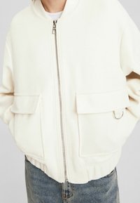 Biała kurtka bomberka o gładkiej fakturze, z metalowym zamkiem błyskawicznym z przodu, dwoma kieszeniami po bokach oraz metalowym akcentem w postaci pierścienia; zestawiona z dżinsami.