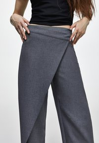 Pantalon gris à jambes larges avec un design unique en croisé à la taille. Le tissu présente une finition lisse, texturée et une apparence sophistiquée.