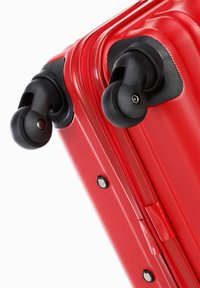 Close-up of red hard-shell suitcase corner with two black spinner wheels and a red zipper against white background.