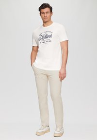 Herren T-Shirt mit kurzen Ärmeln in Cremefarbe und navyblauem Text; kombiniert mit hellbeigen Hosen und Sneakers mit grauen und cremefarbenen Akzenten.
