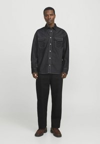 Camicia in denim scuro con due tasche frontali, chiusure a bottoni, cuciture in contrasto, abbinata a pantaloni neri e scarpe marroni. Sfondo neutro.