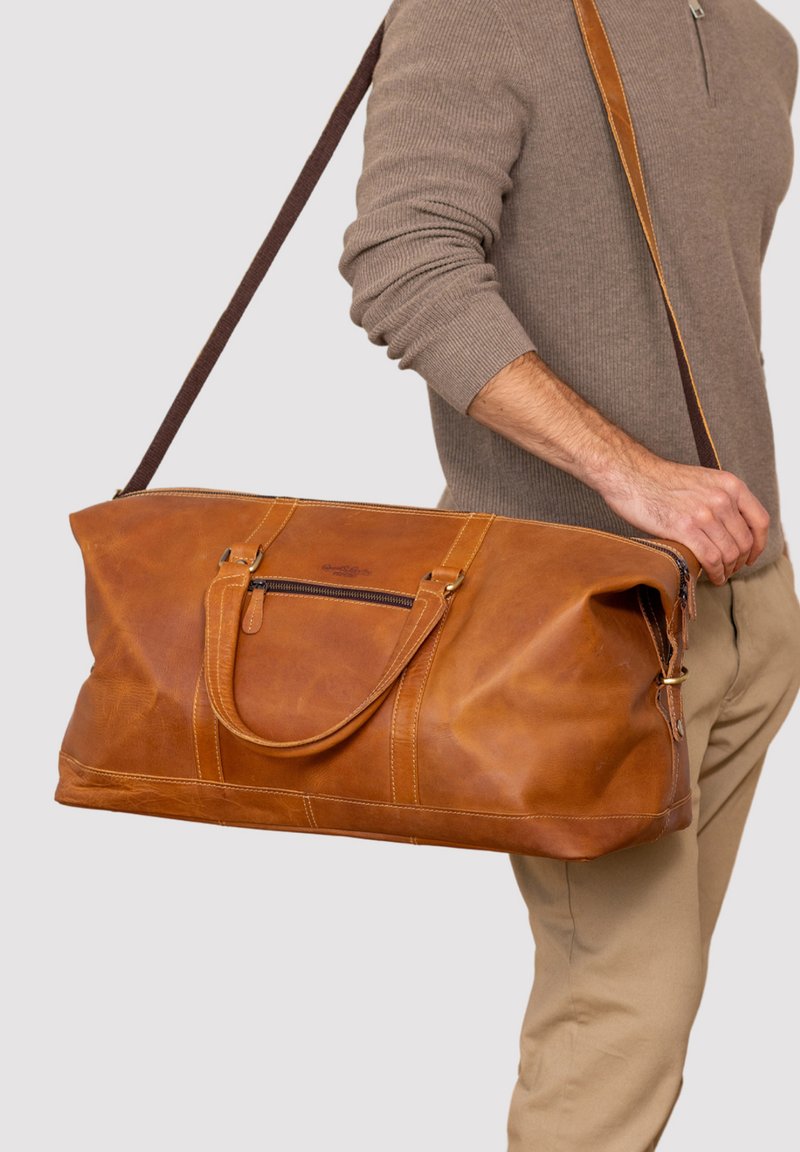 Bruine leren sporttas met dubbele handvatten, verstelbare schouderriem, voorzak met rits en zichtbare stiksels.