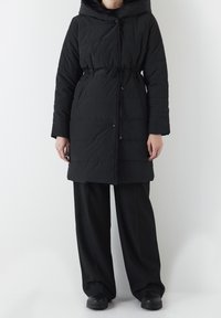 Abrigo de plumas negro con capucha, cintura ajustable con cordón y bolsillos frontales. Textura suave con diseño acolchado y cierres de botones.