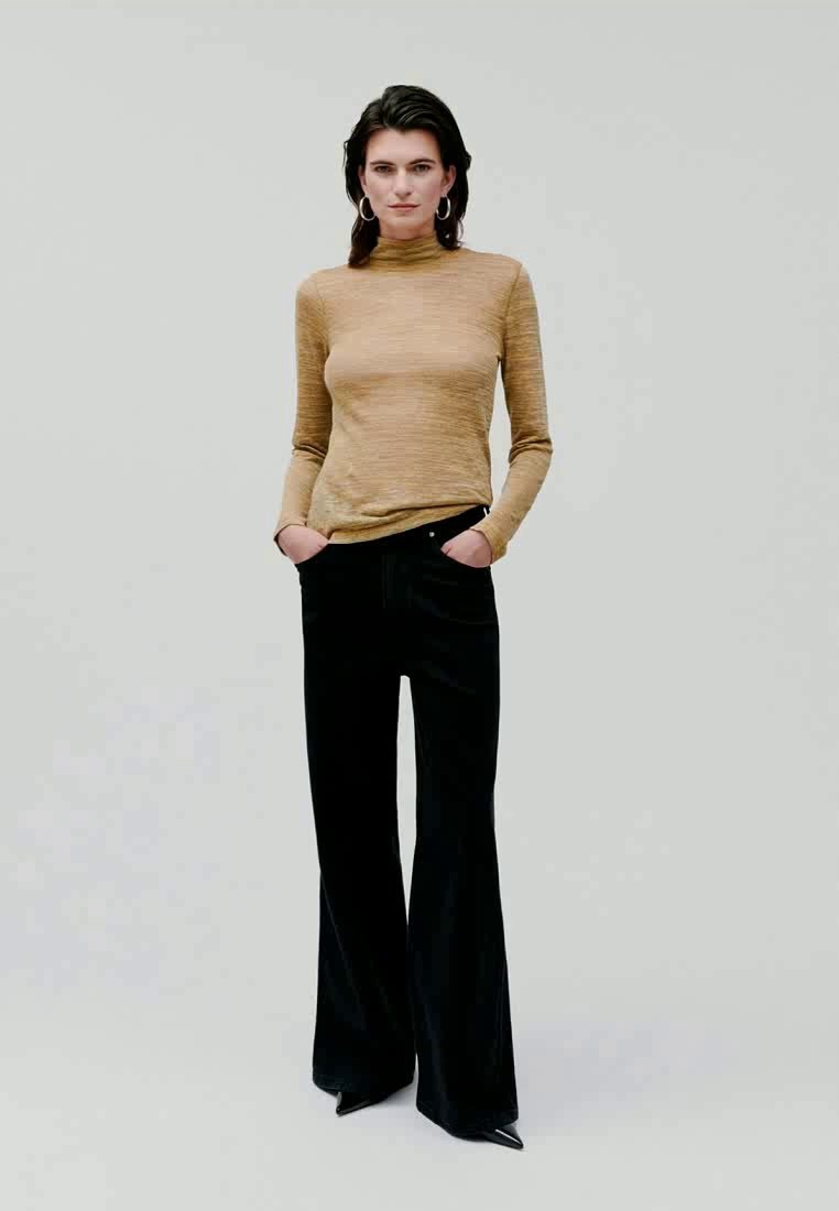 T-shirt ajusté à manches longues en beige doré, associé à un pantalon noir évasé taille haute. Le modèle a les mains dans les poches et porte des boucles d'oreilles créoles.