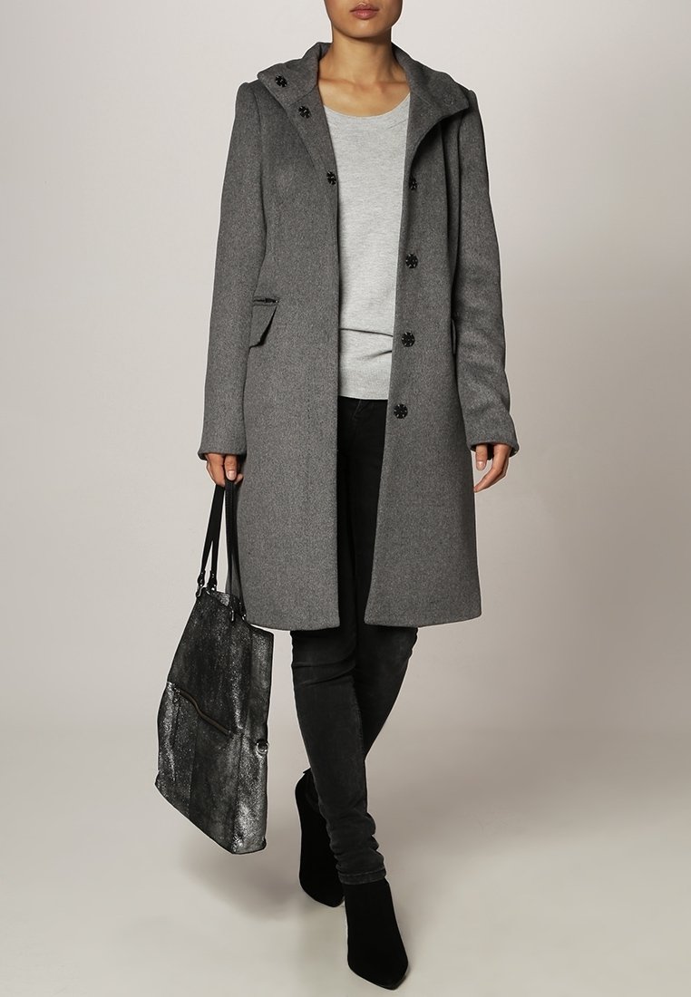 Manteau en laine gris avec capuche, boutons noirs, poches latérales, associé à un haut gris et un jean noir. Tenant un sac métallique et portant des bottes noires.