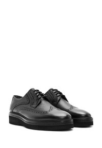 Schwarze Lederbrogues mit durchbrochener Detailgestaltung und dicken Gummisohlen, gezeigt in einer seitlichen Frontalansicht nebeneinander.
