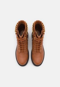 Bottes en cuir marron avec un détail de chaîne dorée autour du col, dotées de lacets, d'œillets métalliques, et d'une semelle en caoutchouc noire.