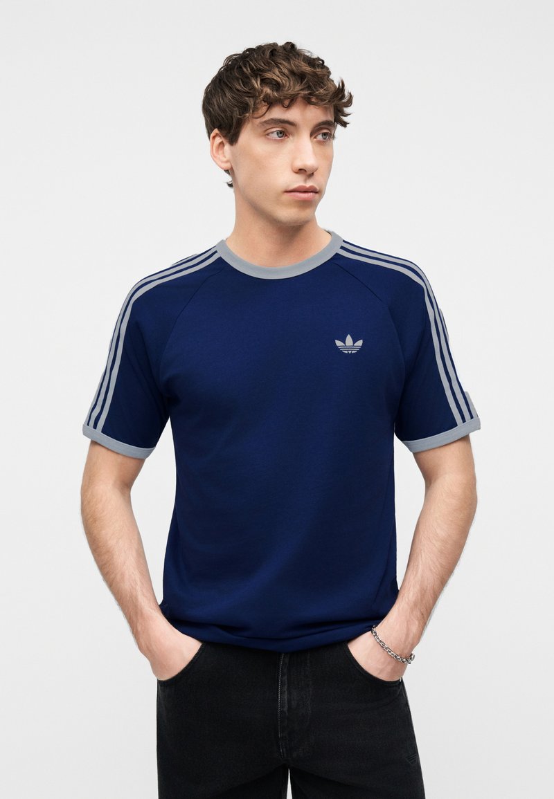 Giovane uomo con capelli corti e ricci indossa una T-shirt Adidas blu navy con rifiniture grigie e strisce, mani nelle tasche dei pantaloni neri.