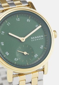 Montre Skagen avec un cadran vert mat, des index et aiguilles dorés, et un bracelet en acier inoxydable argenté et doré.