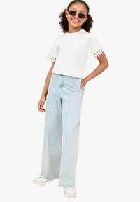 Hvid kortærmet top med blonder, lyse vaskede brede jeans og hvide sneakers. Model iført sorte solbriller og guld halskæde.