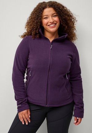 Veste en polaire violette avec un col haut, fermeture éclair à l'avant et poches latérales. Texture douce et design ajusté. Portée sur un pantalon noir.