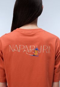 Orange cotton t-shirt featuring a graphic with the word "NAPAPURI" and an illustration of a skier on the back. Short sleeves, crew neck.