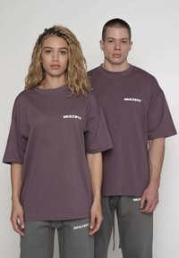 Lilla oversize t-shirts med "MULTIPLY" logo i hvid, kombineret med grå joggingbukser med samme logo på venstre lår.