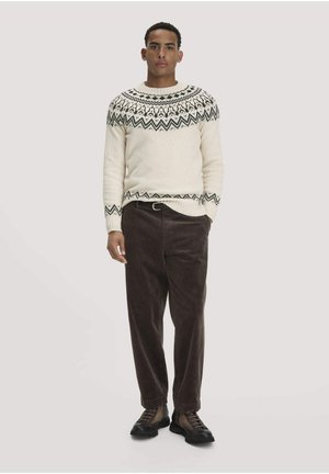 Mann steht und trägt einen cremefarbenen Pullover mit schwarzem geometrischem Yoke-Muster, dunkelbraune Cordhosen und braune Schnürschuhe