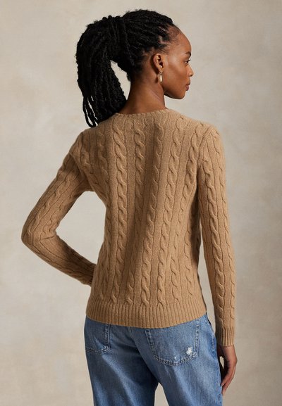 Femme avec une queue de cheval tressée foncée portant un pull en maille torsadée couleur sable et un jean bleu délavé, de dos sur un fond uni.