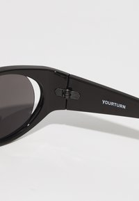 Mustat aurinkolasit tummilla linsseillä, suorakulmainen muoto, kiiltävä muovikehys, saranatiedot ja "YOURTURN" -logo temppeleissä.