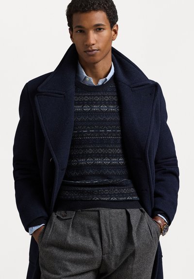 Navy wool overcoat with a wide collar, layered over a dark patterned sweater and light blue collared shirt, paired with grey pleated trousers.