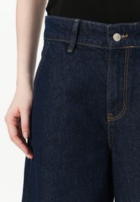 Sötétkék farmernadrág magas derékkal, narancssárga varrással, elülső zsebbel és ezüst gombos záródással. Simaszövet és egyenes lábvágás jellemzi.