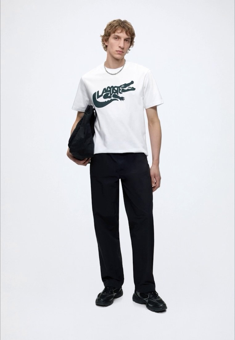 Млад мъж, облечен с бяла тениска с логото на Lacoste, черен панталон, черни маратонки, държащ черна чанта с неутрално изражение на лицето на едноцветен фон.
