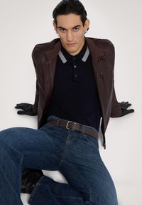 Giacca di pelle marrone con zip, polo blu navy e jeans blu. Guanti testurizzati completano l'outfit. Il modello è seduto con le gambe incrociate.