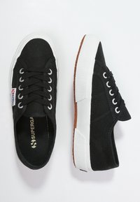 Zapatillas de lona negras con suelas de goma blancas, cordones redondos, tela texturizada y un acento de logo rojo y azul en el lateral.