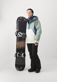Planche de snowboard avec un dessus à motifs noirs, tenue par une personne en veste à capuche vert menthe et blanche, pantalon noir et bottes noires.