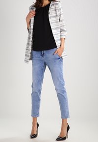 Cardigan ouvert à rayures blanches et noires porté sur un haut noir, associé à un jean bleu clair et des chaussures à talons noirs.