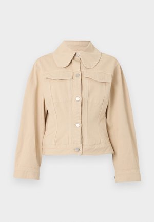 Žena stojí pred jednofarebným pozadím, oblečená v béžovom saku, bielom tričku, voľných zelených nohaviciach a hnedých sandáloch na remienky, ruky má vreckách.