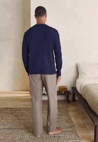 Navy long-sleeve top and beige plaid pajama pants. Smooth fabric, relaxed fit, and lightweight texture. Barefoot on a woven rug.