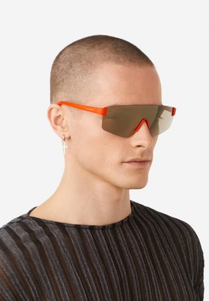 Oranžid päikeseprillid suure ja nurga all disainiga, pruunide poolläbipaistvate läätsedega. Omaduseks on minimalistlik raam ja ninatugi.