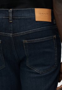 Σκούρο μπλε τζιν με δερμάτινη ετικέτα GANT στη μέση, διακοσμημένο με αντίθετες πορτοκαλί ραφές και δύο πίσω τσέπες.