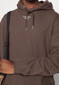 Sudadera marrón con cordones ajustables y extremos con punta de plata. Presenta el logo en blanco "Calvin Klein Jeans" y puños acanalados. Correa negra cruzada en el hombro.