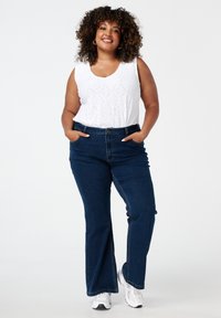 Witte mouwloze top met oogjesdetails, gecombineerd met donkerblauwe flare jeans. Model staat met handen in de zakken en draagt witte sneakers.