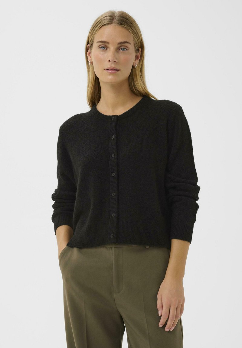 Frau mit blonden Haaren, die eine schwarze Knopfleiste-Strickjacke und olivgrüne Hose trägt, steht mit einer Hand in der Tasche vor einem schlichten Hintergrund.