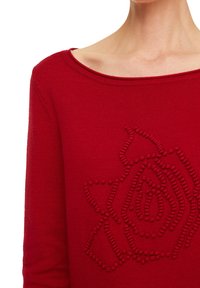 Pull en tricot rouge avec un design de rose texturé à l'avant. L'encolure est large, avec des détails en bord-côte.