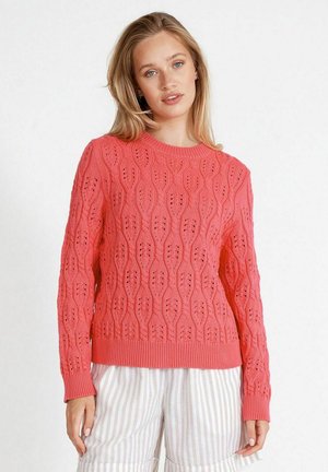 Coral knit sweater with a textured pattern featuring oval cutouts, round neck, and long sleeves, paired with light striped pants.