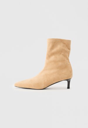 Stivale alla caviglia in suede beige con punta affusolata e tacco kitten nero basso su sfondo bianco semplice.