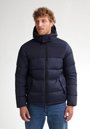 Marineblå polstret jakke med høj krave og lynlåslukning, der har sidelommer og en tekstureret overflade. Båret over blå jeans.