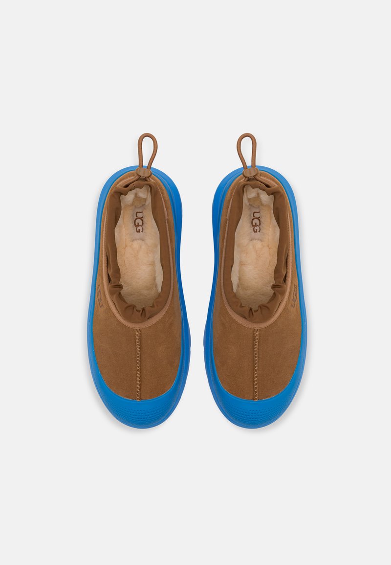 Zapatos slip-on de ante marrón con suelas de goma azul, que cuentan con un forro suave y un puño elástico para un ajuste cómodo. Logo de UGG impreso en el interior.