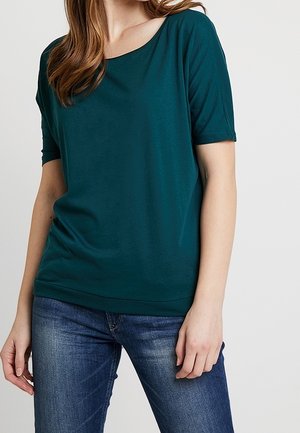 Femme portant une chemise manches courtes bleu sarcelle foncé et un jean bleu, debout avec une main détendue le long du corps.