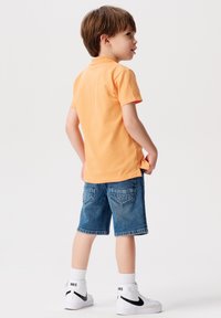 Oranje polo shirt met korte mouwen en een kraag, gecombineerd met blauwe denim shorts. Witte sneakers met zwarte accenten en geribde witte sokken.