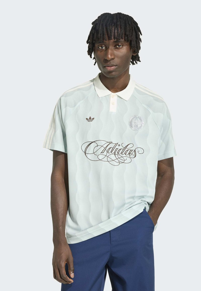 Mladý muž s krátkými dreadlocky, který má na sobě bílé polo tričko Adidas s nápisovým logem a tmavě modré kalhoty, stojící před jednobarevným pozadím.