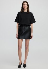 Zwart oversized t-shirt gecombineerd met een zwarte leren minirok, voorzien van een gedefinieerde taille en subtiele glans, aangevuld met zwarte hakken.