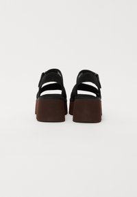 Zwarte platform sandalen met brede bruine blokhakken en enkelbandjes, van achteren weergegeven tegen een witte achtergrond.