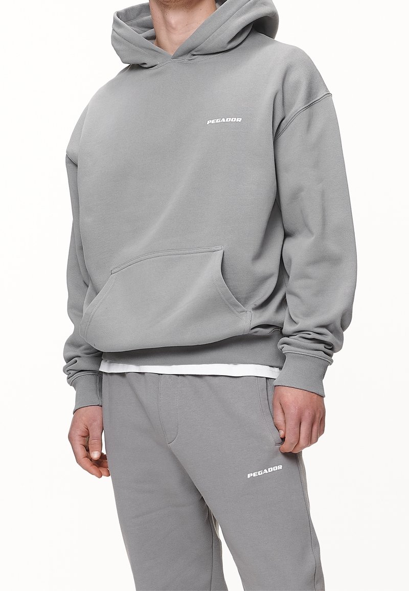 Felpa grigia con cappuccio, tasca a marsupio, polsini e orlo a coste; presenta il logo bianco "PEGADOR" sul petto e pantaloni grigi abbinati.